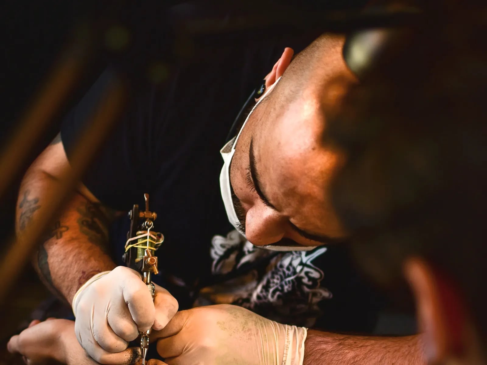 A tattoo artist leaning in to work on a client’s forearm.